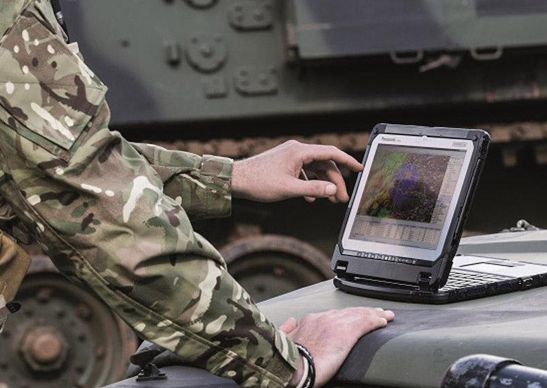 Person in camo jacket using a tablet while a tank is in the backgroud