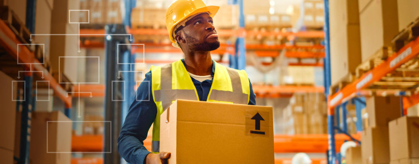 Man with safety hat and high visibility vest holding a box looking up into shelves.